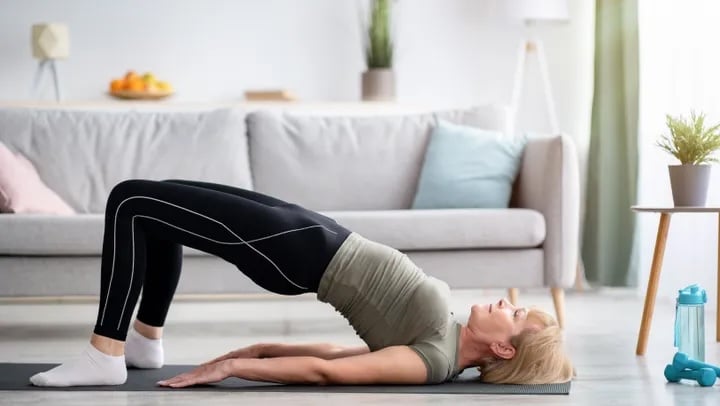 Person performs a glute bridge exercise on a yoga mat in a bright living room. The movement targets the glutes by lifting the hips off the floor while keeping the shoulders and feet grounded, making it a popular exercise for building lower body strength.