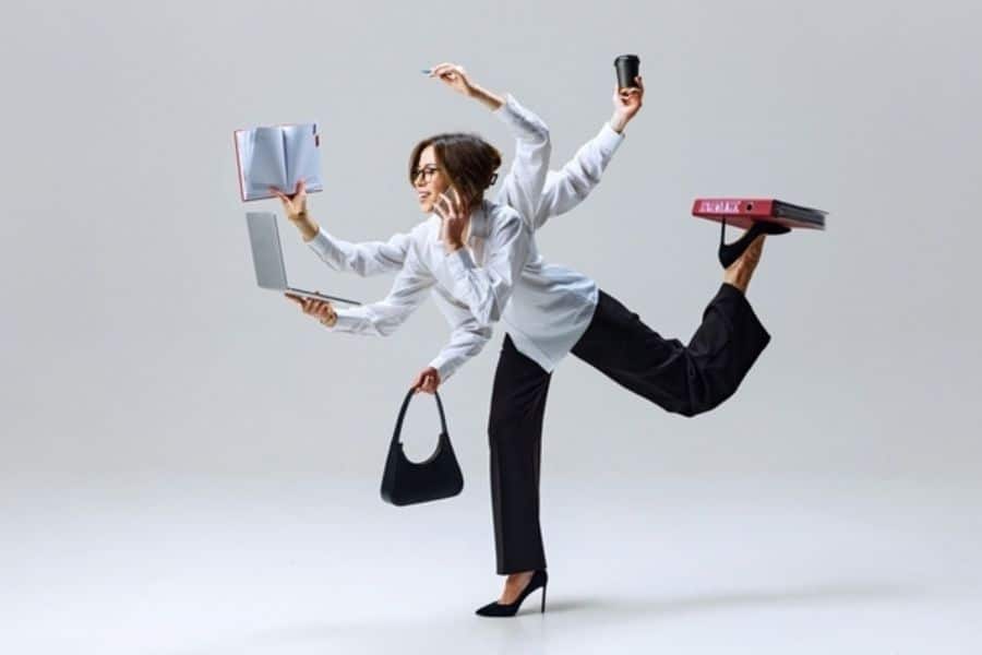 A businesswoman balances on one leg while holding a laptop handbag notebook coffee cup and office folder with several arms. The playful scene represents multitasking and the mental strain of managing many responsibilities at once.
