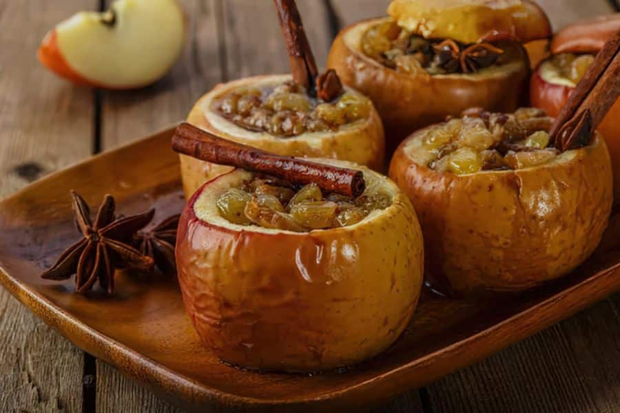 Baked apples filled with oats and raisins on a wooden plate with cinnamon sticks and star anise. A warm fruit based snack option for a PCOS friendly recipe roundup.