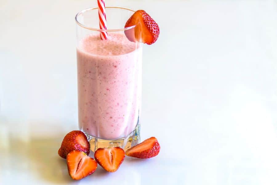 Pink strawberry smoothie in a tall glass with a striped straw and fresh strawberry slices around it. A simple fruit smoothie for a PCOS friendly snack idea.