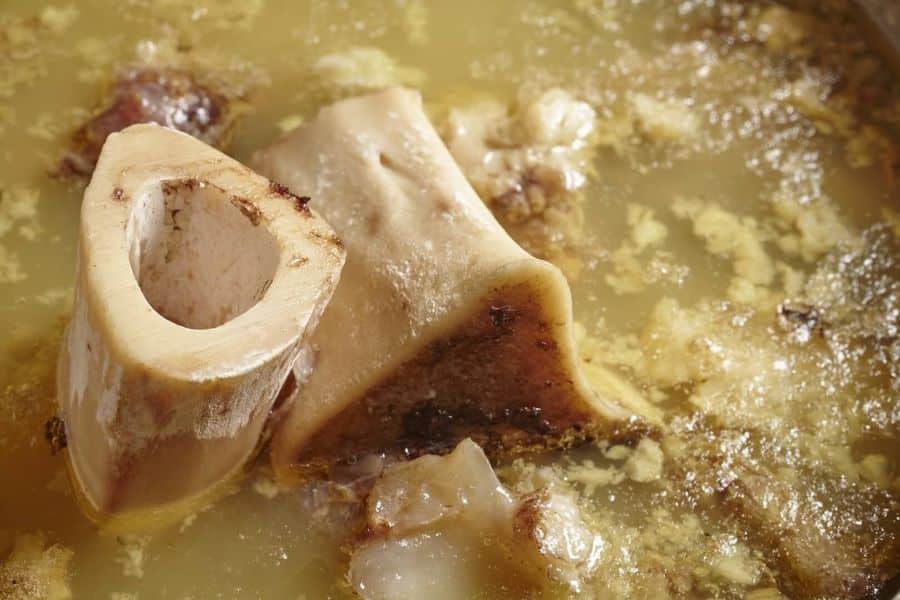 Large marrow bones and pieces of meat simmer in a cloudy golden broth with beads of fat across the surface. The close view highlights the rich texture of homemade bone broth.