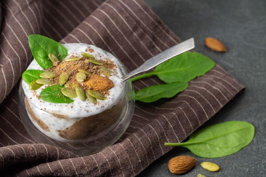Small glass of chia pudding topped with yogurt. pumpkin seeds. almonds. spinach leaves. and cinnamon. The snack is set on a brown striped cloth with extra almonds and greens nearby.
