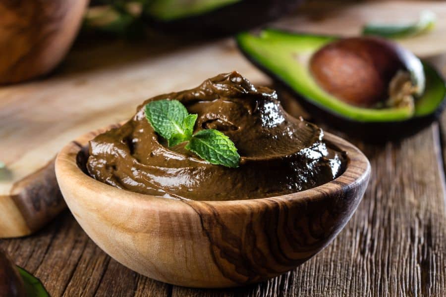 Creamy chocolate avocado pudding in a wooden bowl with fresh mint on top. A halved avocado sits in the background.
