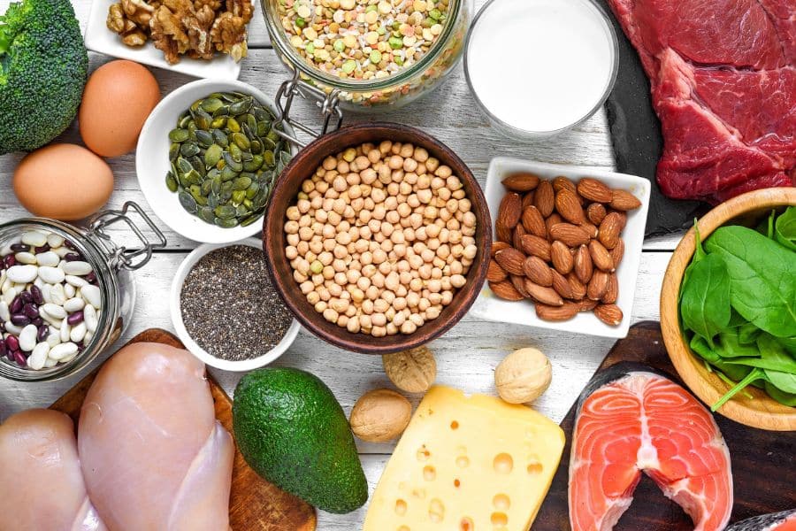 Assortment of protein rich foods arranged on a wooden table including chicken breast, salmon, eggs, chickpeas, beans, almonds, cheese, milk, lentils, seeds, spinach, avocado, broccoli, and walnuts. The variety highlights both animal and plant based sources of protein for a balanced diet.