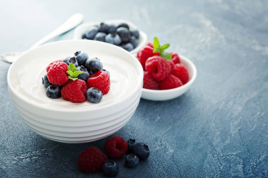 Bowl of creamy yogurt topped with blueberries. raspberries. and mint. Small bowls of extra berries sit behind it on a blue surface.