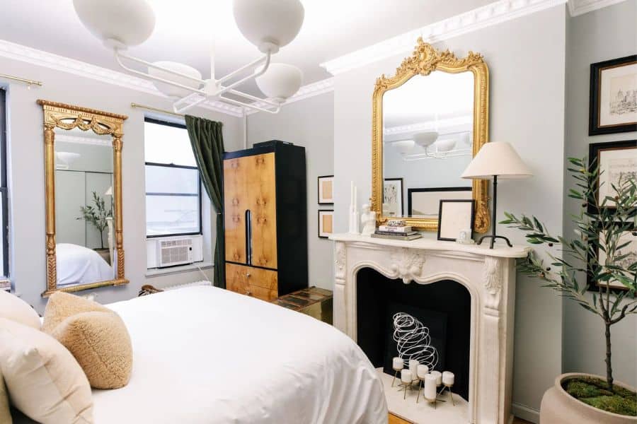 A bright bedroom with white bedding sage green walls and two ornate gold mirrors. One mirror hangs above a white fireplace mantel and another hangs beside the bed near a black framed window.