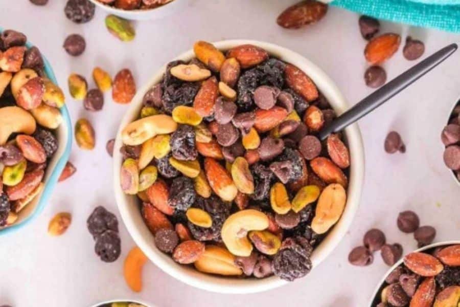 Bowl of trail mix with almonds. cashews. pistachios. dried fruit. and chocolate chips. A spoon rests in the bowl for a quick PCOS friendly snack.