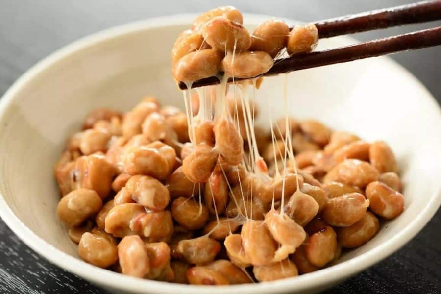 Wooden chopsticks lift sticky fermented soybeans from a white bowl of natto. Long translucent strands stretch between the glossy beans and the bowl.