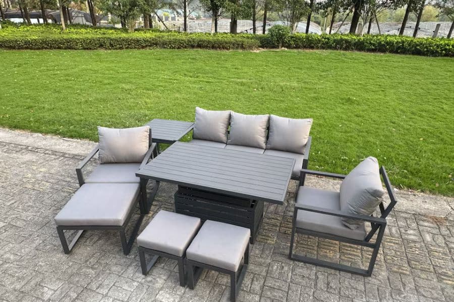 A gray outdoor sectional dining set with cushioned seats surrounds a square table on a stone patio. A green lawn and hedges fill the background.