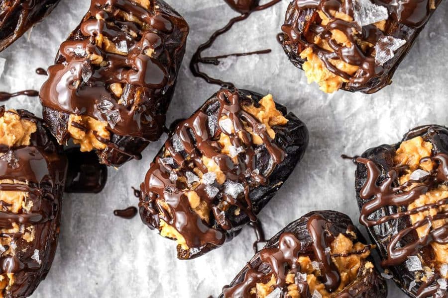 Dark dates filled with creamy nut butter and drizzled with melted chocolate on parchment paper. Flaky sea salt is scattered on top for a sweet PCOS friendly snack idea.