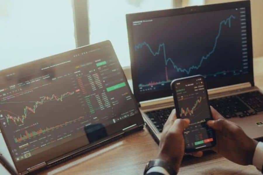 Person holding a smartphone displaying stock market charts while monitoring investing performance across two laptops with market data and price graphs on screen. The setup shows active investing and tracking of market movements in real time.