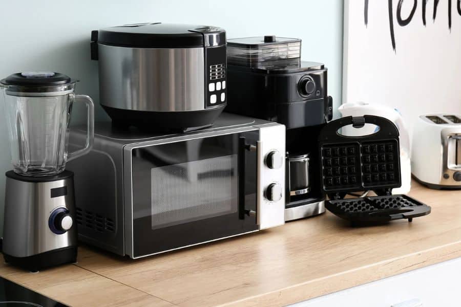 A countertop setup with a blender microwave multicooker coffee maker waffle maker and toaster arranged against a pale wall. Part of the wall art reads "Home."
