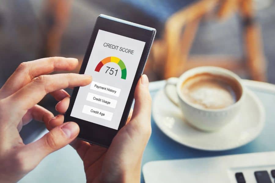 Person checks a phone displaying "CREDIT SCORE" and "751" beside a color gauge, with options for "Payment History" "Credit Usage" and "Credit Age." A cup of coffee and laptop sit nearby, suggesting a casual review of credit score details.