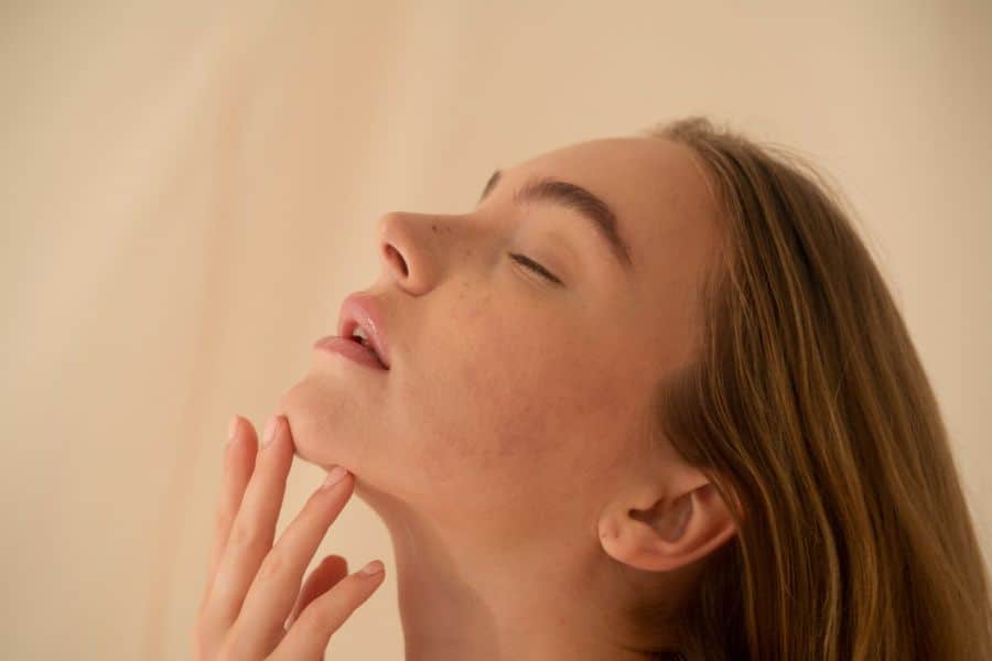 A woman tilts her head back with her eyes closed and gently touches the area beneath her chin. The close profile view draws attention to the jawline and double chin area.