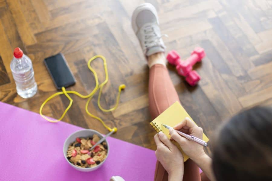 A woman sits beside a purple exercise mat and writes in a yellow notebook after a workout. A bowl of fruit and cereal sits nearby with dumbbells and a water bottle as she plans how to keep the weight off.