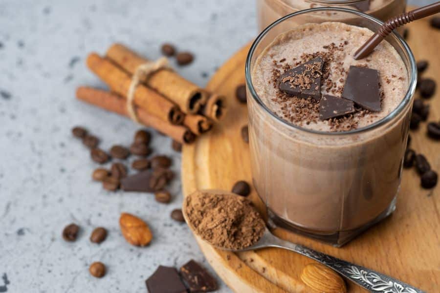 Chocolate smoothie in a glass topped with dark chocolate squares and shaved chocolate. Cinnamon sticks. coffee beans. cocoa powder. almonds. and chocolate pieces are arranged around it.