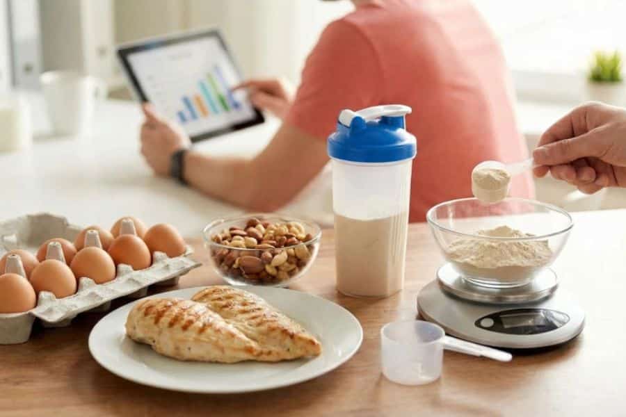 Person measures protein powder into a bowl on a kitchen scale beside grilled chicken, eggs, mixed nuts, and a prepared shake. A tablet displaying charts in the background suggests tracking nutrition and protein intake.