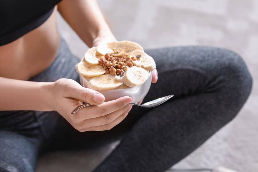Person in workout clothes holds a bowl topped with banana slices and walnuts while sitting after exercise. The snack combines protein and nutrient dense ingredients that support recovery and satiety.