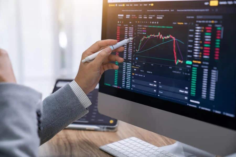 Close up of a hand pointing at a computer monitor displaying stock charts, price movements, and market indicators during an investing analysis session. The screen highlights changing market conditions and investment research.