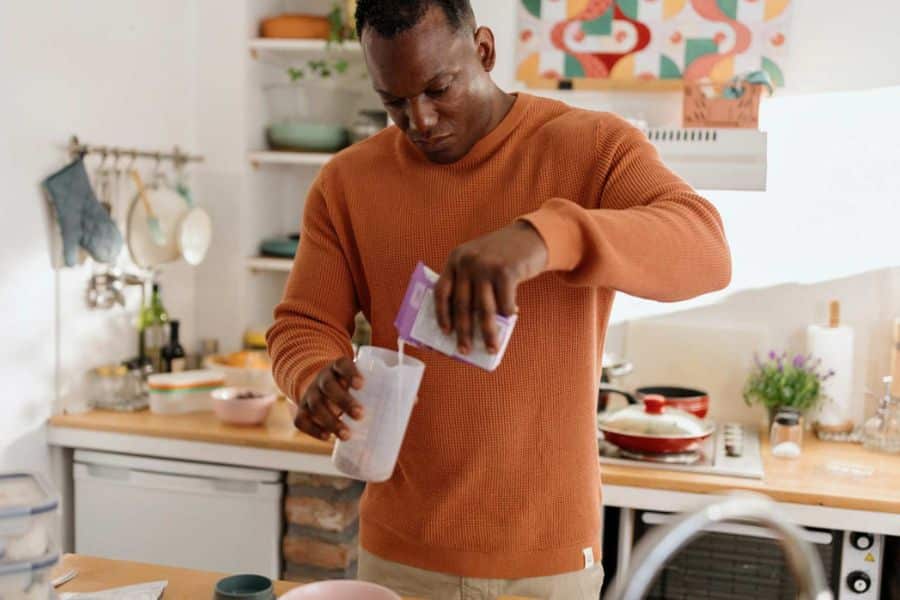 Man prepares a protein shake in a home kitchen by pouring powdered supplement into a shaker bottle. The scene illustrates a convenient way to add protein to a daily nutrition routine.