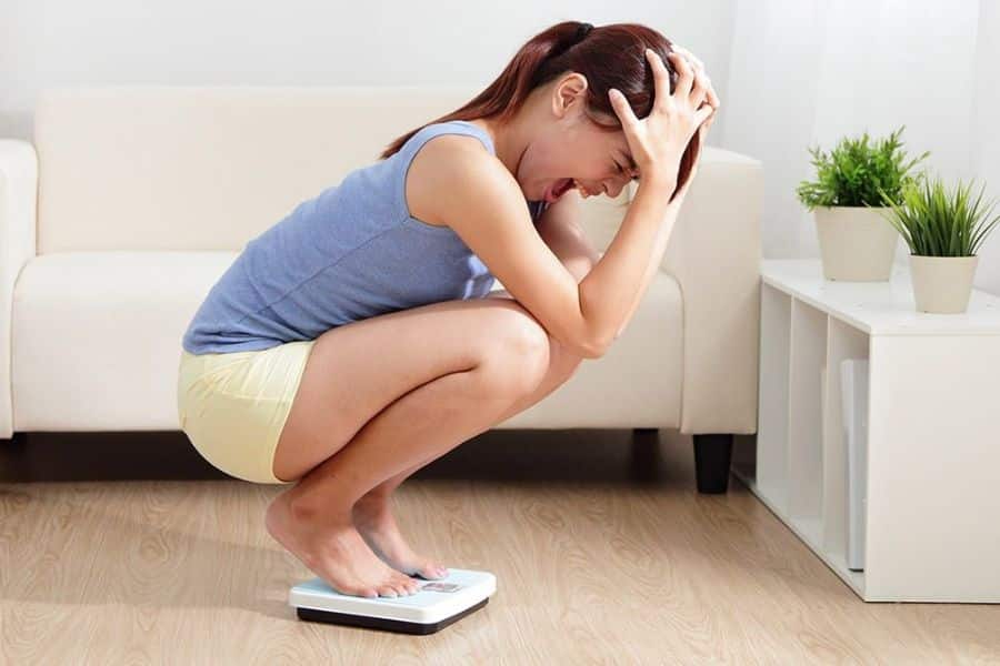 A distressed woman crouches on a bathroom scale with both hands gripping her head. Her tense posture shows frustration or disappointment about her weight.
