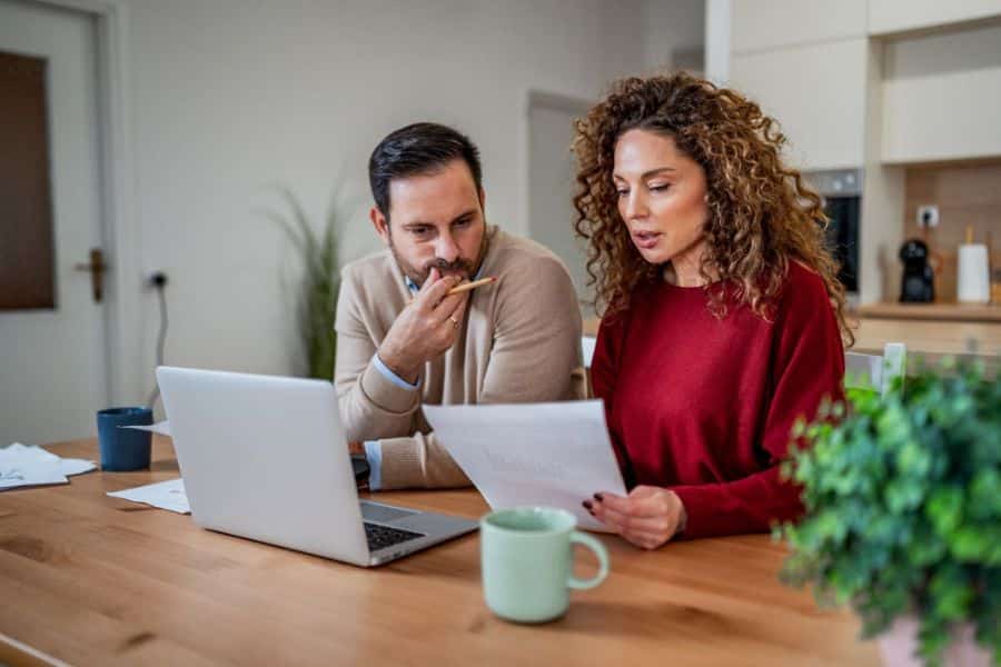 Couple reviewing financial documents at a kitchen table with a laptop while discussing investing goals and household finances. The scene highlights planning investment decisions and managing long term financial objectives.