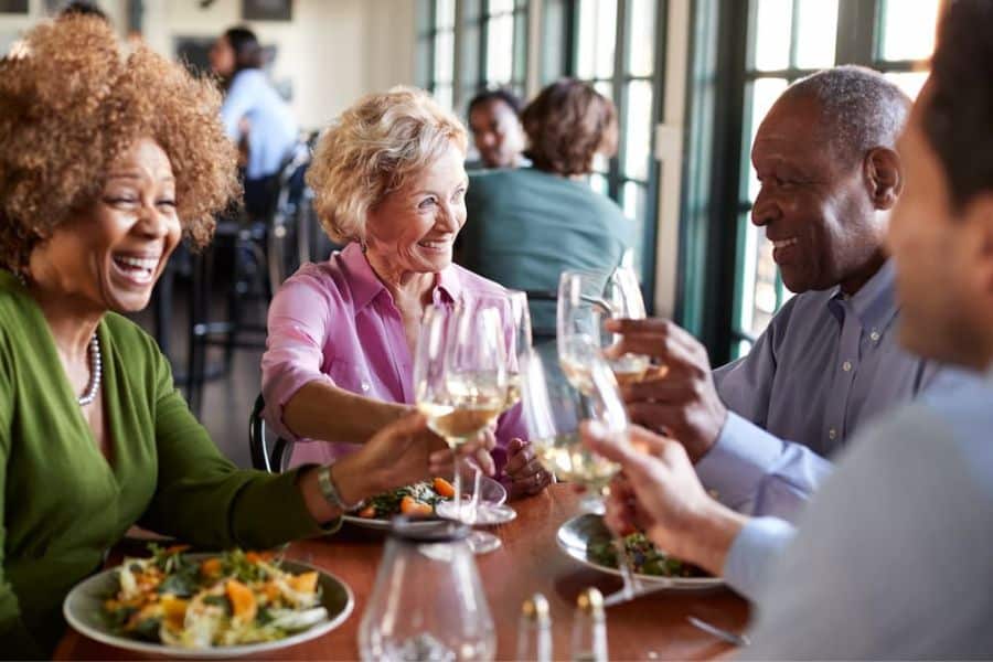 Four older adults share a meal and raise wine glasses together at a restaurant. Their laughter and relaxed conversation suggest an enjoyable social outing.