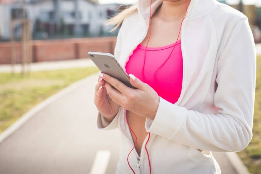 Woman in a white jacket and pink sports top holds a phone while wearing earbuds outdoors. The scene suggests using music or a fitness app to support walking habits.