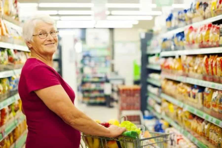 An older woman pushes a cart filled with fresh produce through a grocery store aisle. She smiles toward the camera while shopping for food.