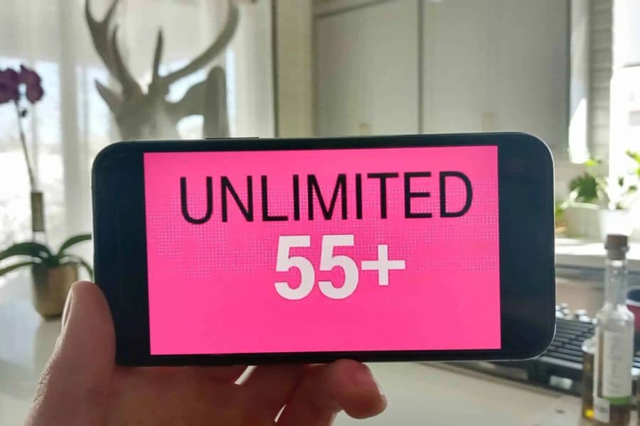 A hand holds a smartphone displaying the words "UNLIMITED 55 PLUS" in black and white lettering on a bright pink background. The offer appears designed for customers aged 55 and older.