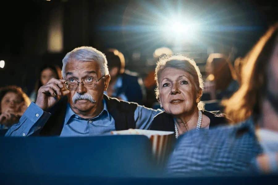 An older couple watches a movie together in a dark theater with a tub of popcorn in front of them. The man adjusts his glasses while both focus on the screen.