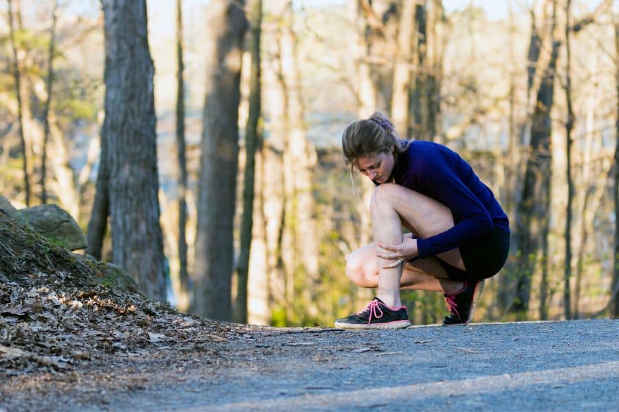 Runner crouches on a wooded trail while holding her lower leg. The scene shows how pain or strain can interrupt walking habits and outdoor exercise.
