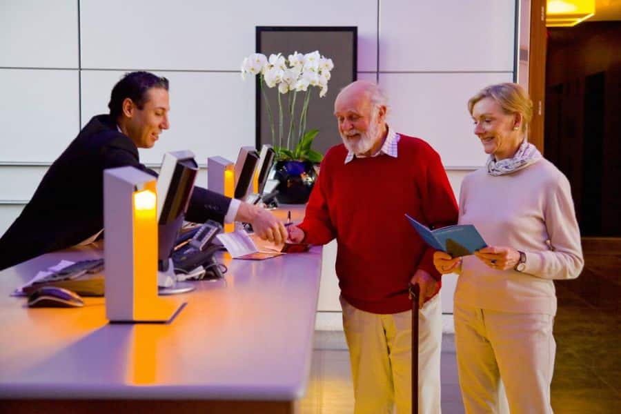 An older couple checks in at a hotel reception desk while a staff member hands them paperwork. The man holds a cane and the woman carries an open travel document.