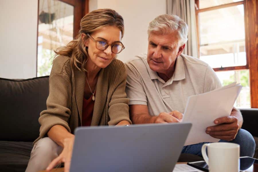 An older couple reviews paperwork beside an open laptop at home. They appear to be comparing bills finances or available discounts together.