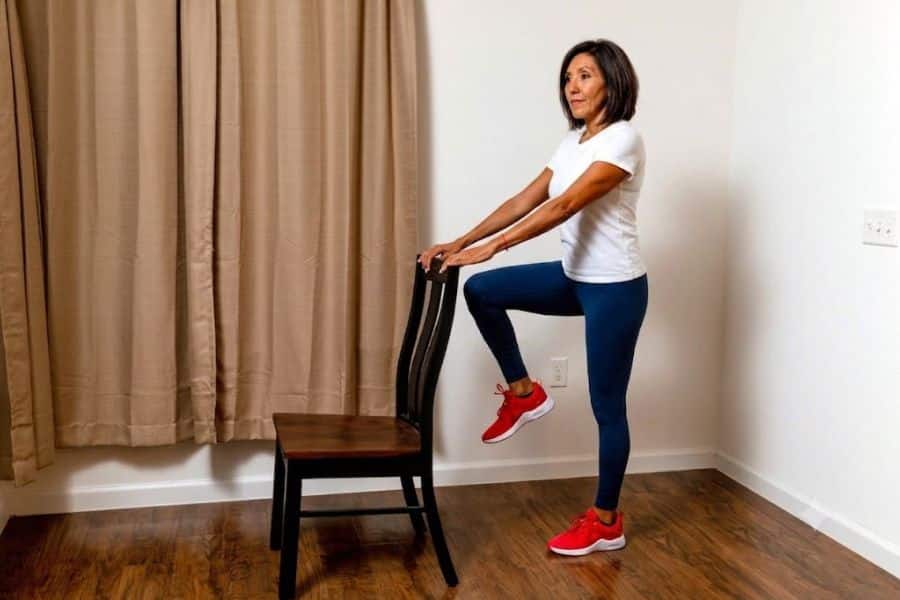 A woman practices a standing knee raise at home while holding the back of a wooden chair for support. The movement demonstrates a supported variation of the 10 second balance test.