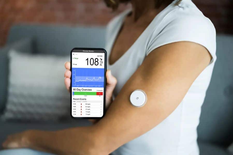 Person wearing a continuous glucose monitor on their upper arm while holding a smartphone showing a glucose reading of "108 mg dL". The close up connects type 2 diabetes management with tracking blood sugar patterns throughout the day.