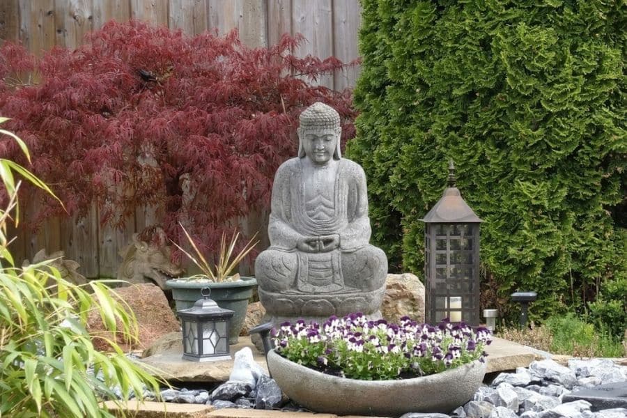 Stone Buddha statue seated behind a bowl of purple and white flowers. Red Japanese maple leaves evergreen shrubs rocks and black lanterns create a calm meditation garden.