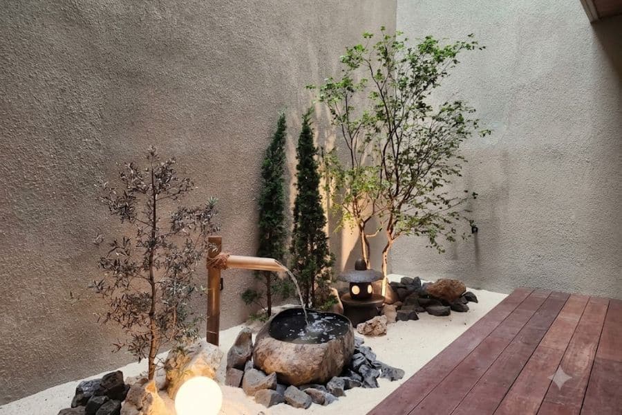 Minimal Japanese courtyard with a bamboo water spout flowing into a round stone basin. White gravel dark rocks slender trees and a glowing lantern frame a wooden deck designed as a Japanese style retreat.