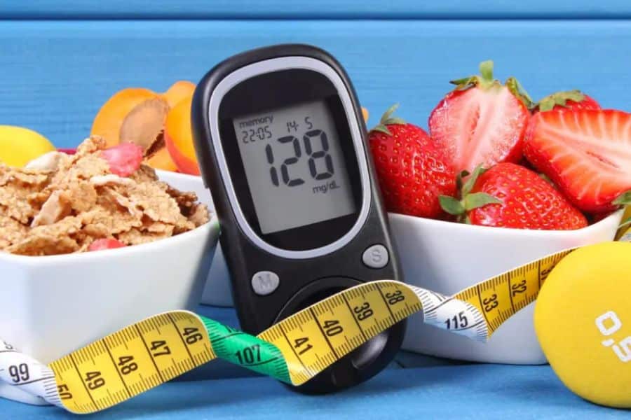 Blood glucose meter showing "128 mg dL" beside strawberries. cereal. sliced fruit. a measuring tape. and a yellow dumbbell. The setup suggests type 2 diabetes management through food choices. glucose monitoring. and exercise.