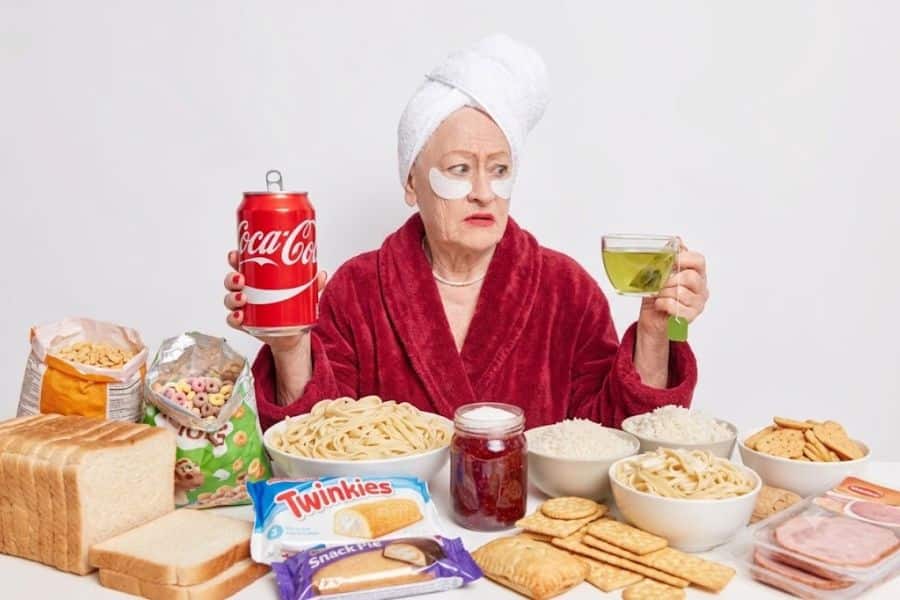 Older woman in a red robe holding a can of Coca Cola and a cup of green tea while sitting in front of white bread. sugary cereal. pasta. rice. crackers. cookies. lunch meat. jam. Twinkies. and a SnackWell’s package. The spread highlights processed and high sugar foods that age the body and skin.