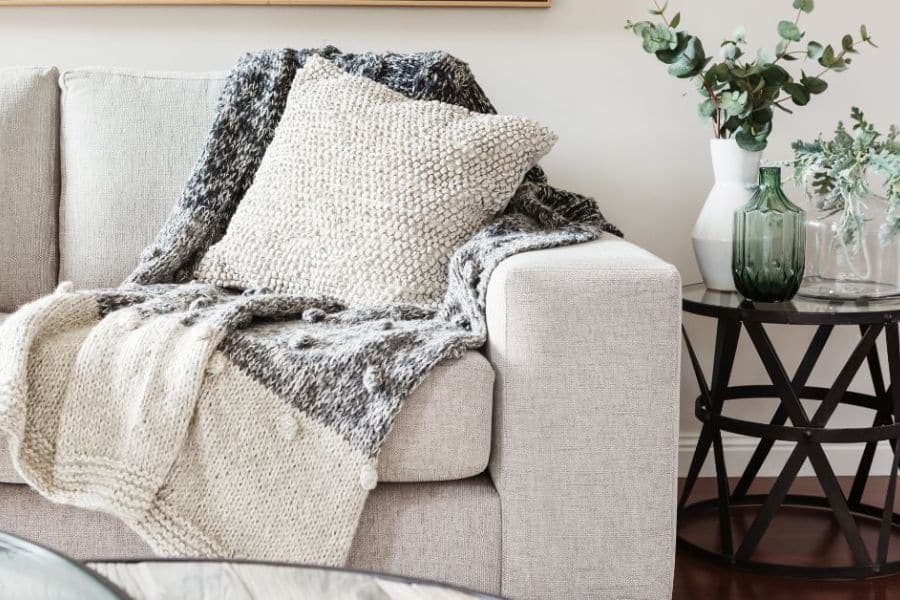 Close up of a neutral sofa styled with layered knit blankets. a textured cream pillow. and a side table with vases and greenery for a cozy staged home detail.