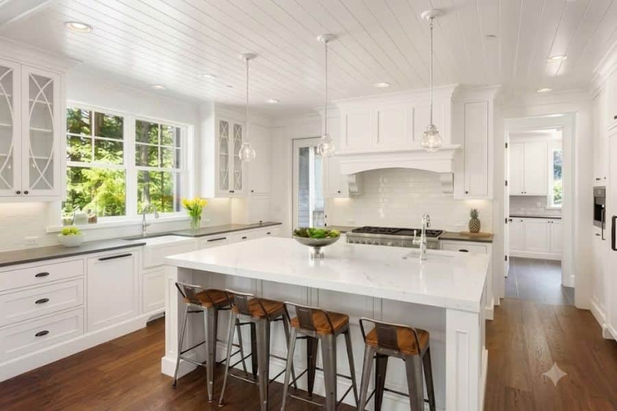 White kitchen staged with a large marble island. wood stools. glass pendant lights. fresh flowers. and greenery to make the room feel bright. clean. and welcoming.
