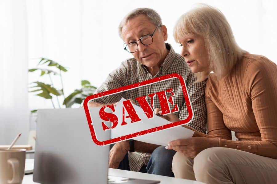 Older couple reviewing paperwork together on a laptop with a large red "SAVE" stamp over the image, representing ways to save money on Medicare costs. The image includes the text "SAVE."