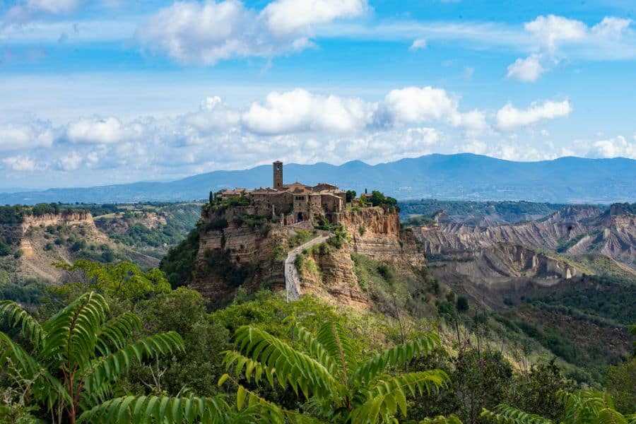 Historic hilltop village perched on a rocky cliff above a rugged green valley. Mountains stretch across the horizon beneath a bright blue sky filled with clouds.
