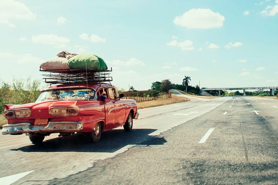 Vintage red car loaded with luggage drives along a wide open highway beneath a bright blue sky. The road stretches toward the horizon suggesting an independent road trip.