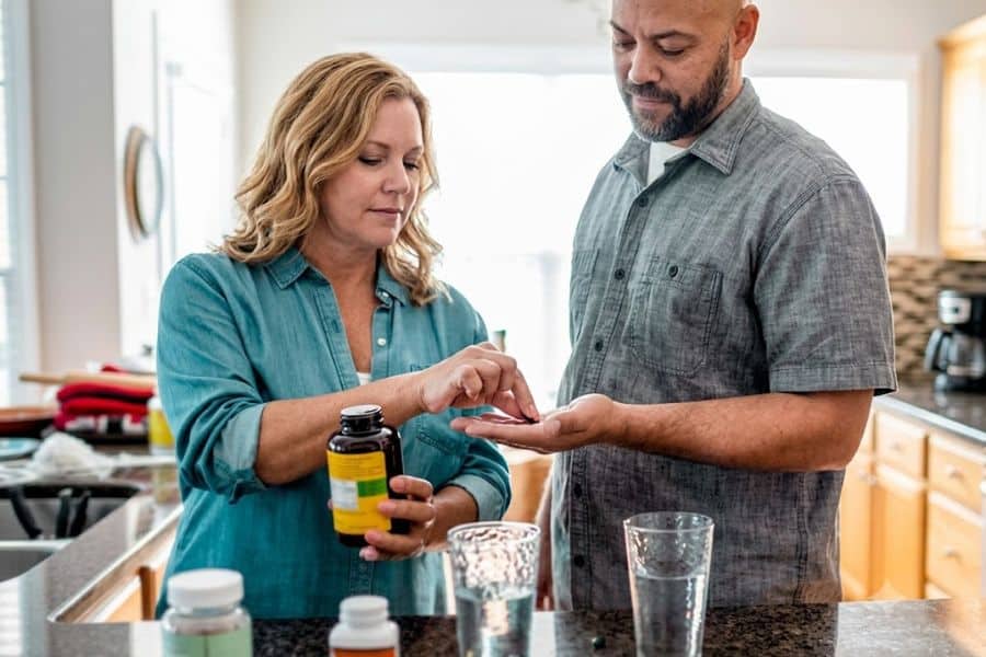 Adult couple stands together in a kitchen as one person hands vitamins to the other from a supplement bottle beside glasses of water. The scene supports guidance on when to take your vitamins as part of a daily health routine.