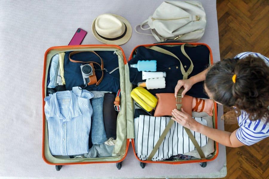 Traveler packs an open suitcase with folded clothing camera toiletries and small bags while securing items with luggage straps. A hat phone and backpack sit nearby as the traveler prepares for a trip.