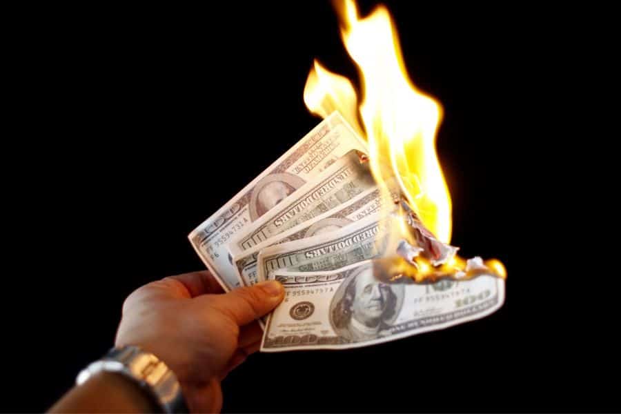 A hand holds several burning one hundred dollar bills against a black background as bright flames consume the money.