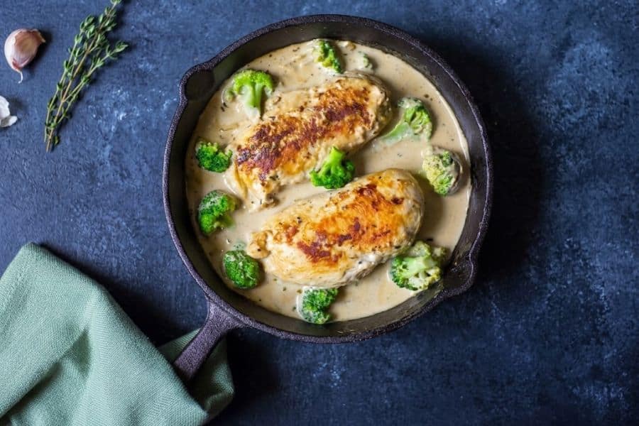Two browned chicken breasts simmer in a creamy sauce with bright green broccoli inside a cast iron skillet. Fresh thyme and garlic sit beside the pan.