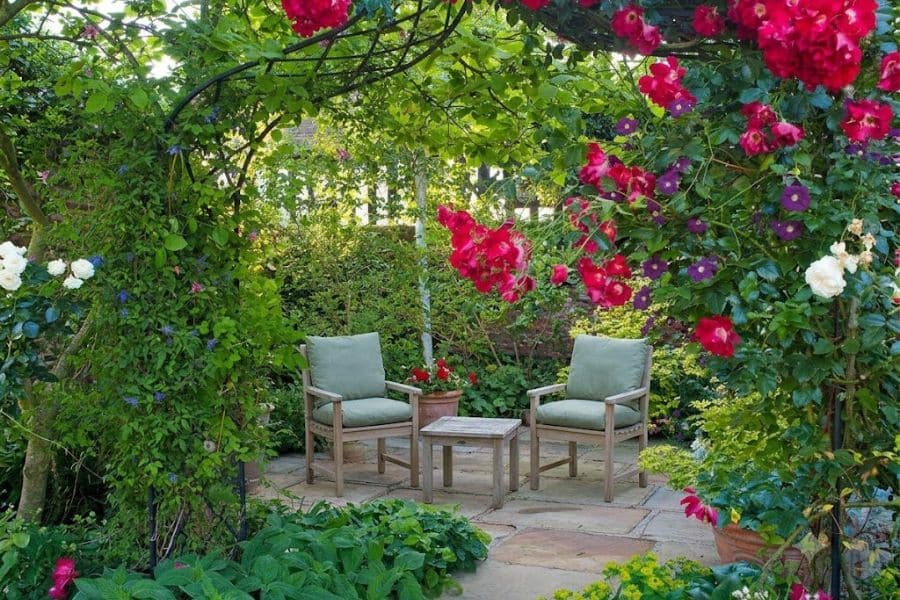 Two cushioned chairs and a small wooden table sit beneath an arch covered in red roses and green vines. White and purple flowers surround the secluded garden seating area.